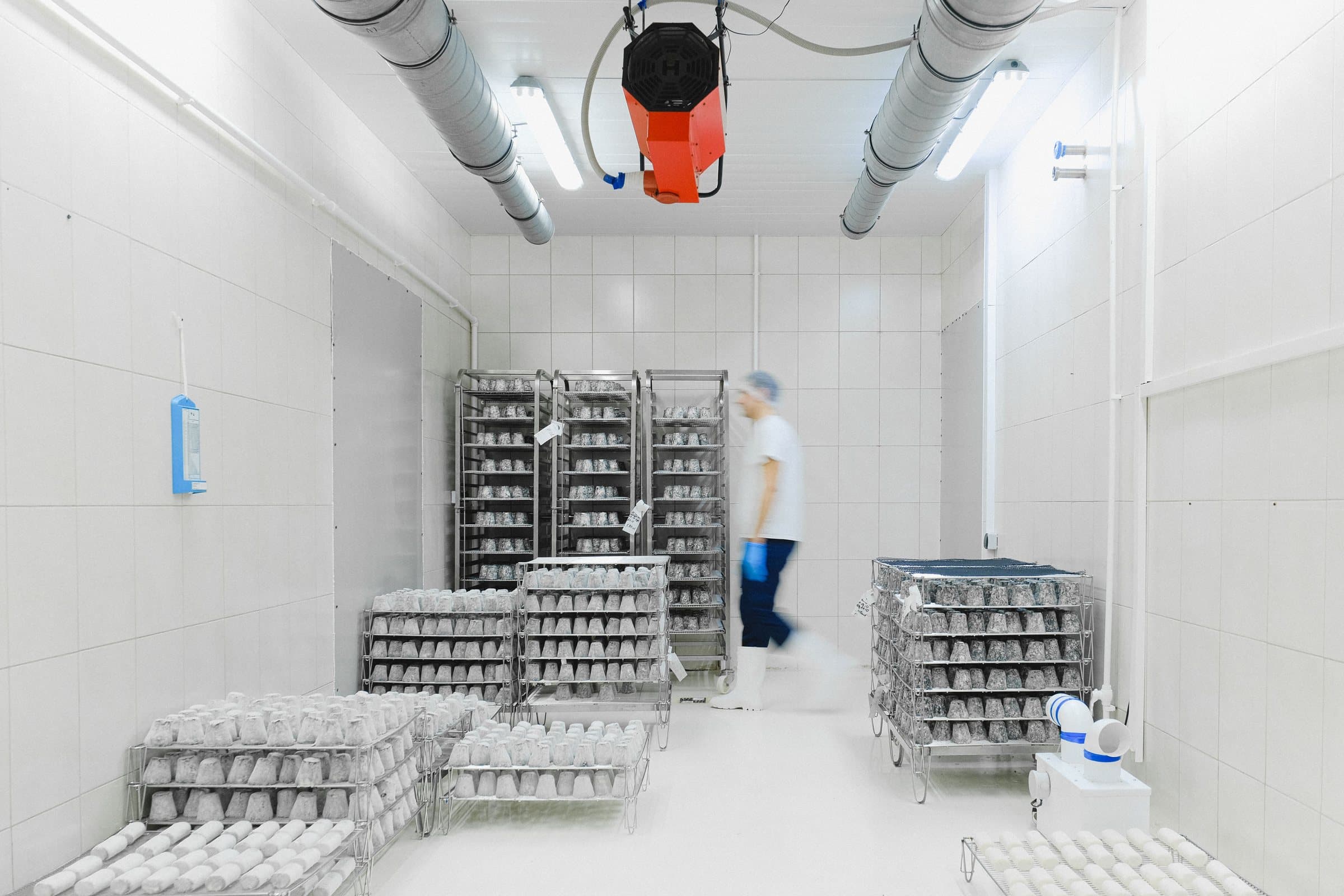 A white-tiled temperature-controlled food storage room with air ducts overhead and steel trolley racks of wrapped product.