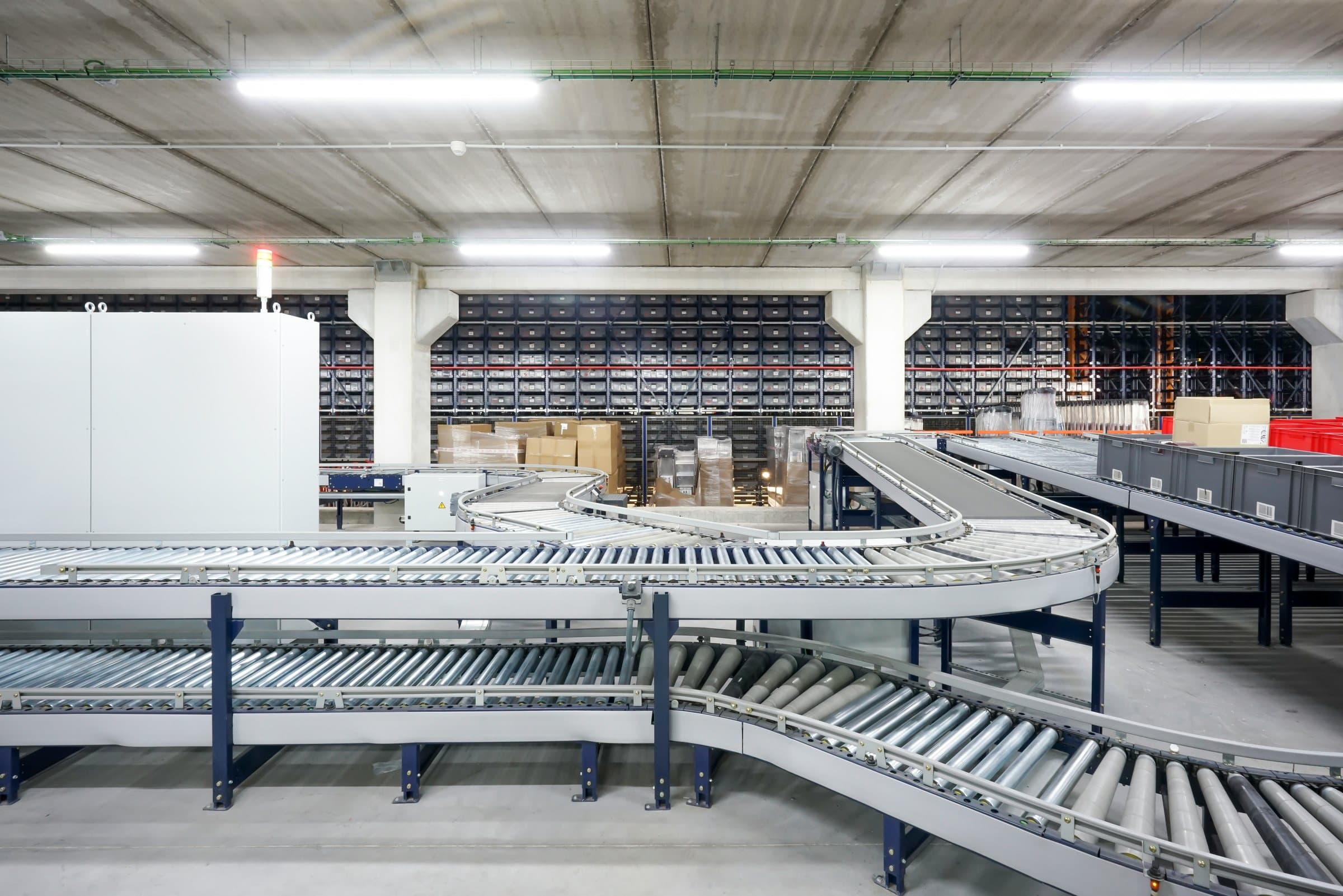 Two levels of curved roller conveyor in a warehouse, with totes on a side line and a wall of storage bins behind.