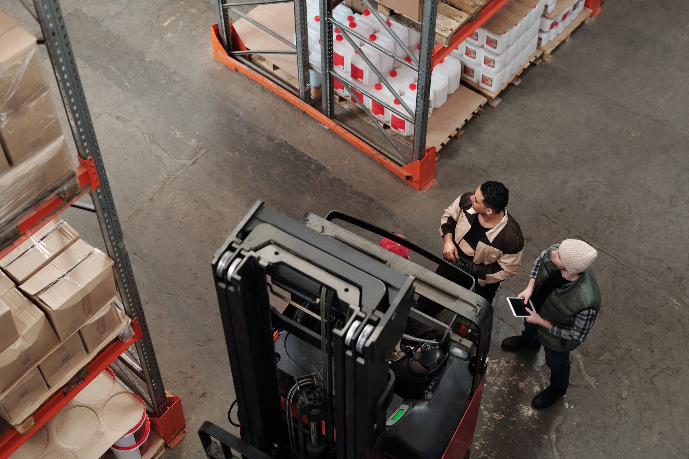 Two warehouse workers, one holding a tablet, beside a forklift between pallet racks, seen from above.